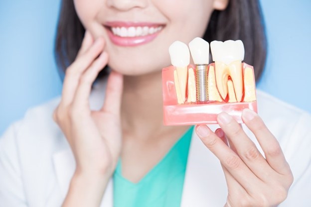 dentist smiling, touching cheek, and holding dental implant model